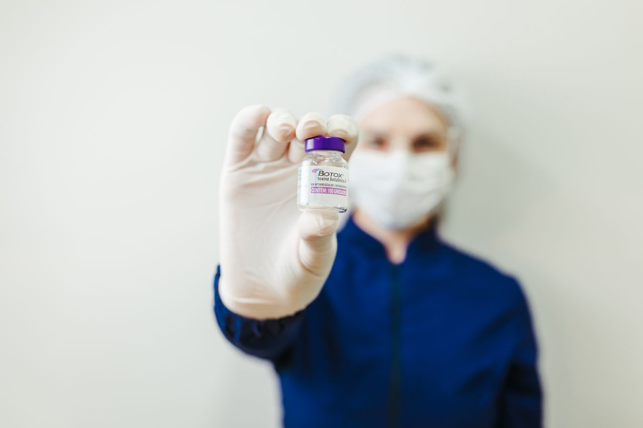 Features Healthcare worker holding a Botox vial in a clinical setting, wearing protective gear.